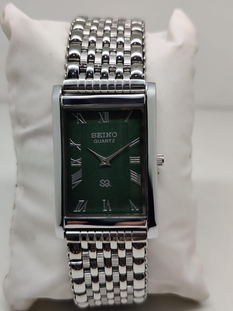 Może przedstawiać: Prostokątny zegarek Seiko Quartz ze srebrną kopertą i bransoletą. Tarcza zegarka jest ciemnozielona ze srebrnymi rzymskimi cyframi i wskaz&oacute;wkami. Bransoleta składa się z małych, połączonych metalowych ogniw.