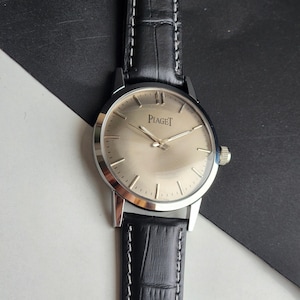 Puede incluir: Un reloj de pulsera Piaget plateado con esfera clara y agujas plateadas. El reloj tiene una correa de cuero negro con costuras blancas. La esfera muestra el nombre de la marca "Piaget".
