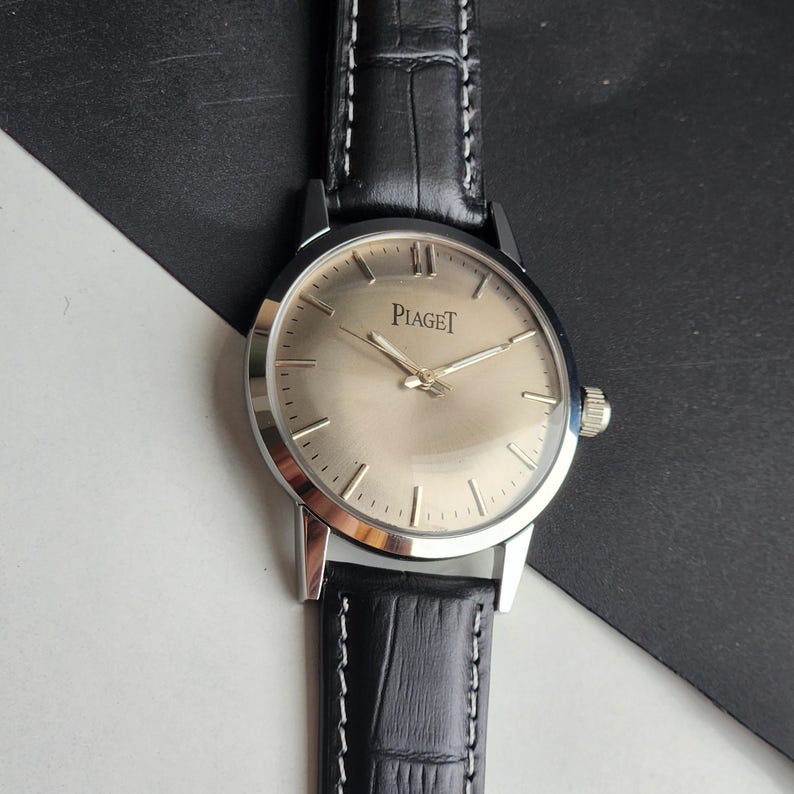 Puede incluir: Un reloj de pulsera Piaget plateado con esfera clara y agujas plateadas. El reloj tiene una correa de cuero negro con costuras blancas. La esfera muestra el nombre de la marca "Piaget".