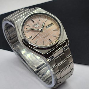 May include: A silver-toned wristwatch with a light pink dial featuring a grid pattern. The watch face displays the day and date. The watch has a silver band and the word "Automatic" is visible on the watch face.