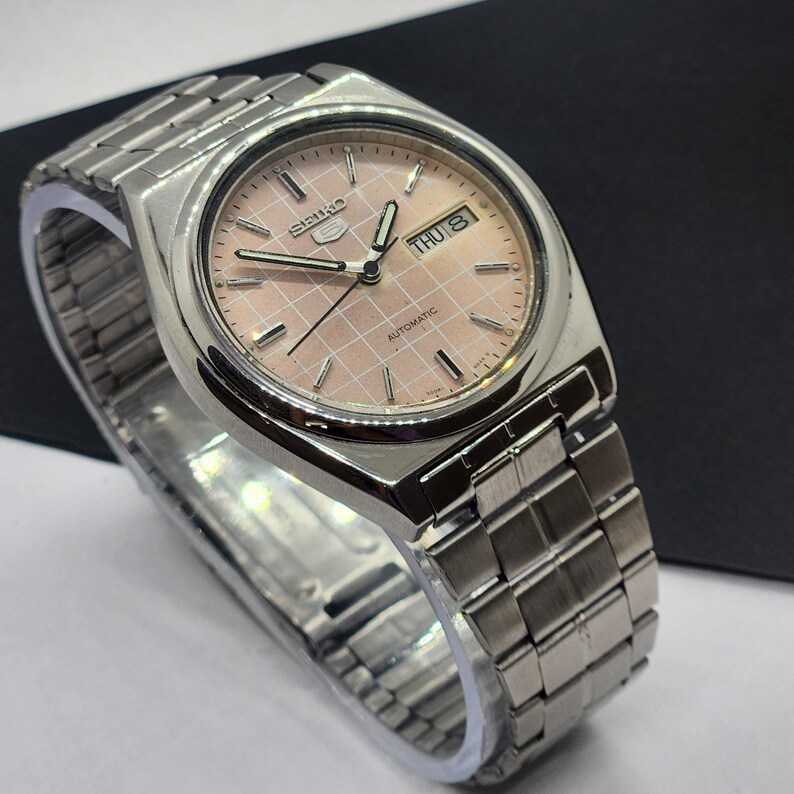 May include: A silver-toned wristwatch with a light pink dial featuring a grid pattern. The watch face displays the day and date. The watch has a silver band and the word "Automatic" is visible on the watch face.