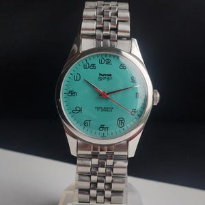 May include: A silver wristwatch with a turquoise face and a silver band. The watch face features red hands, Arabic numerals, and text in a foreign language. The watch is labeled "PARA SHOCK 17 JEWELS" and "INDIA".