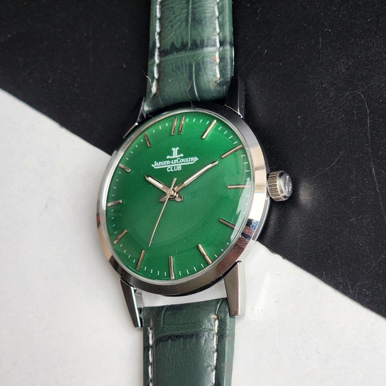 May include: A round wristwatch with a green face and a silver-colored case. The watch has a green leather band. The watch face has the words "Jaeger-LeCoultre CLUB" printed on it. The watch hands are silver-colored.