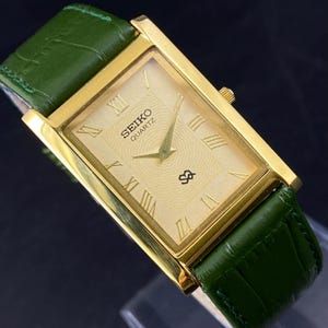 May include: A gold-toned Seiko Quartz watch with a rectangular face and Roman numeral hour markers. The watch features a textured dial and is paired with a green leather strap. The watch case is approximately 1 inch wide.