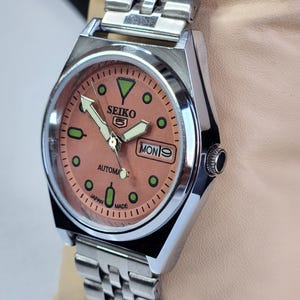 K&ouml;nnte beinhalten: Eine Seiko 5 Automatik-Uhr aus Edelstahl mit einem lachsfarbenen Zifferblatt. Die Uhr verf&uuml;gt &uuml;ber eine Tages- und Datumsanzeige, Leuchtzeiger und gr&uuml;ne Markierungen. Das Armband ist silberfarben.