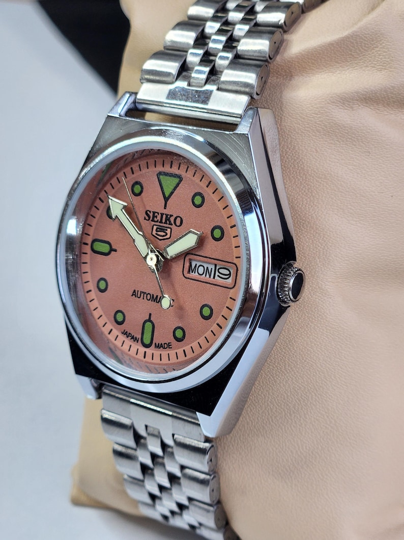 K&ouml;nnte beinhalten: Eine Seiko 5 Automatik-Uhr aus Edelstahl mit einem lachsfarbenen Zifferblatt. Die Uhr verf&uuml;gt &uuml;ber eine Tages- und Datumsanzeige, Leuchtzeiger und gr&uuml;ne Markierungen. Das Armband ist silberfarben.