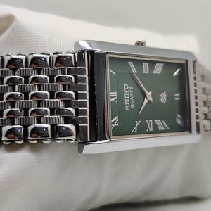 Może przedstawiać: Prostokątny zegarek Seiko Quartz z zieloną tarczą i srebrnymi rzymskimi cyframi. Zegarek ma srebrny metalowy pasek o plecionym wzorze. Tarcza zegarka wyświetla logo Seiko Quartz. Zegarek leży na białej powierzchni.