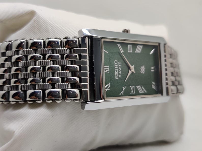 Może przedstawiać: Prostokątny zegarek Seiko Quartz z zieloną tarczą i srebrnymi rzymskimi cyframi. Zegarek ma srebrny metalowy pasek o plecionym wzorze. Tarcza zegarka wyświetla logo Seiko Quartz. Zegarek leży na białej powierzchni.