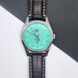 May include: A wristwatch with a teal face and silver casing. The watch has a dark brown leather band. The face displays Hindi numerals and text. The watch hands are red. The watch is a classic style.