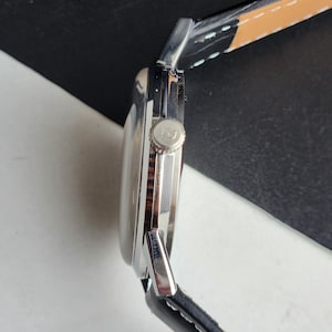 Puede incluir: Un reloj de pulsera plateado con una correa de cuero negro. La esfera del reloj es redonda y la corona es visible en el lateral. La correa es negra con costuras blancas y un acento marr&oacute;n.