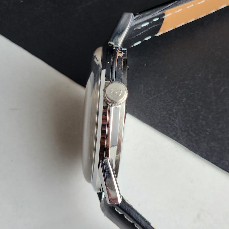 Puede incluir: Un reloj de pulsera plateado con una correa de cuero negro. La esfera del reloj es redonda y la corona es visible en el lateral. La correa es negra con costuras blancas y un acento marr&oacute;n.