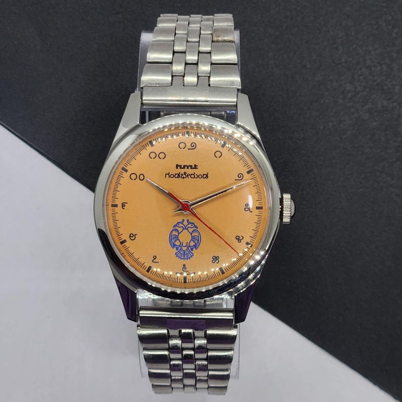 May include: A silver wristwatch with a light orange face and a silver band. The watch face features black numerals, a red second hand, and a blue emblem. The watch has the text "hmt" and another line of text in a foreign language.