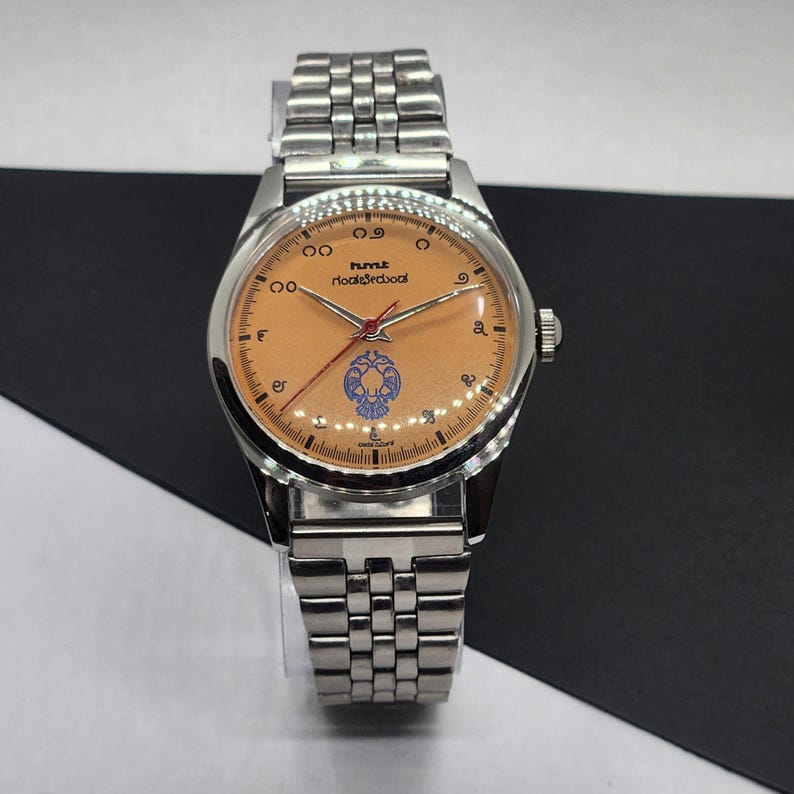 May include: A silver-toned wristwatch with a light orange face and a matching silver band. The watch face features black numerals, a red second hand, and blue and white decorative elements. The watch is displayed on a clear stand.