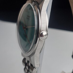 May include: A silver wristwatch with a green face and red hands. The watch has a clear, domed crystal and a silver band. The watch face has the word "TIME" printed on it. The watch is a classic style.