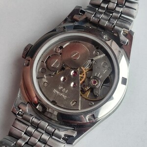 May include: A silver-toned wristwatch with a visible mechanical movement. The watch's back reveals gears, jewels, and intricate internal components. The watchband is made of interlocking metal links. The watch is a vintage style.