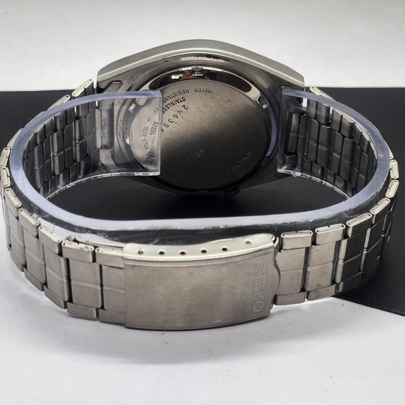 May include: A stainless steel Seiko watch with a silver metal band. The watch face is black with silver text and hands. The back of the watch has the text "Water Resistant Stainless Steel" and the model number 2N639F.