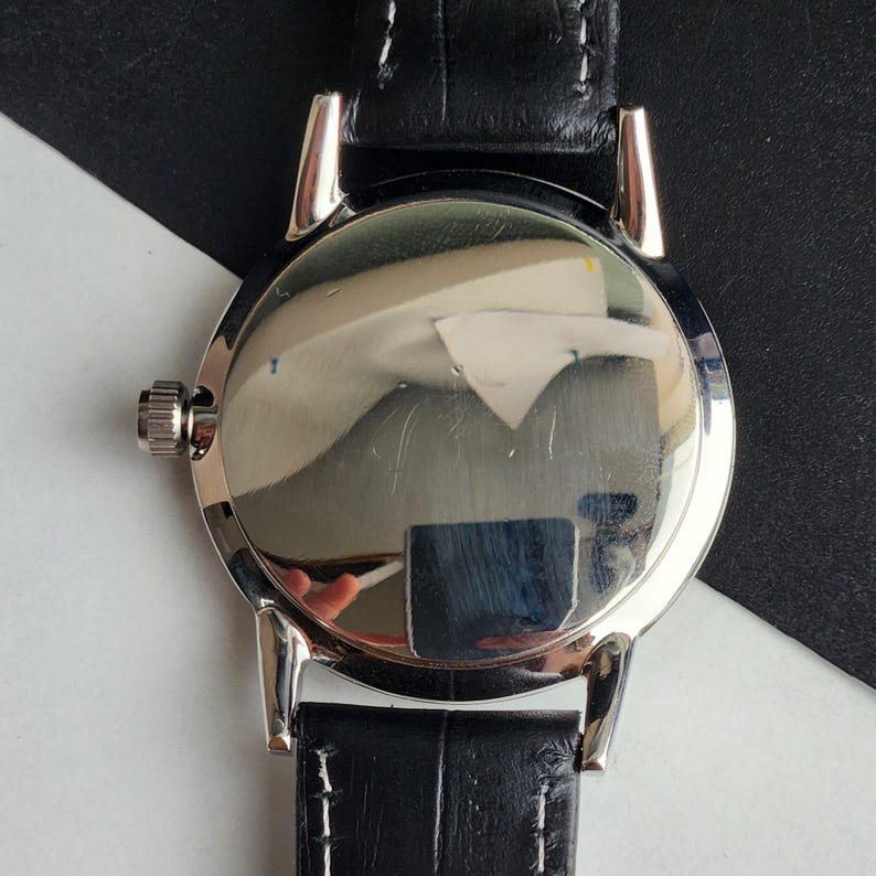 Puede incluir: Un reloj de pulsera plateado con una correa de cuero negro. El reloj tiene una esfera redonda con una superficie reflectante. La correa est&aacute; cosida con hilo blanco. La corona del reloj es visible en el lado izquierdo.
