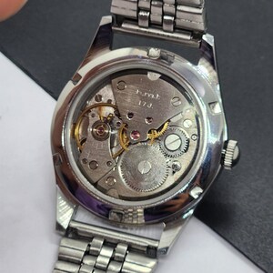 May include: A close-up view of the back of a silver wristwatch, revealing the intricate mechanical movement. The watch's inner workings, including gears, jewels, and the balance wheel, are visible through the clear case back. The watch has a silver metal band.