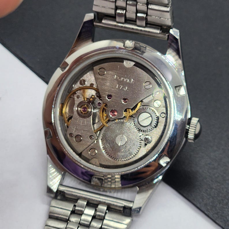 May include: A close-up view of the back of a silver wristwatch, revealing the intricate mechanical movement. The watch's inner workings, including gears, jewels, and the balance wheel, are visible through the clear case back. The watch has a silver metal band.