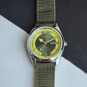 May include: A Citizen automatic wristwatch with a green and yellow face and a green nylon strap. The watch face displays the day and date. The watch has a silver-coloured case.