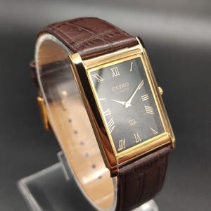 Puede incluir: Un reloj Seiko Quartz rectangular dorado con esfera gris oscuro y números romanos. El reloj tiene una correa de cuero marrón. El reloj se exhibe sobre un soporte transparente.