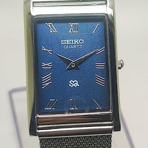 Puede incluir: Un reloj Seiko Quartz rectangular con esfera azul y números romanos plateados. El reloj tiene una correa de malla plateada y una caja plateada. La esfera muestra el logotipo de Seiko y la palabra "QUARTZ".