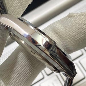 May include: Close-up of a silver wristwatch with a polished metal case. The watch has a sleek, modern design with a reflective surface. The watch is held by a white fabric.