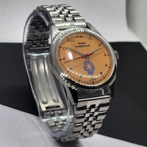 May include: A silver wristwatch with a light orange face and a silver band. The watch face has black numerals, red hands, and blue and white decorative elements. The watch is displayed on a clear stand.
