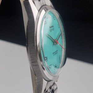 May include: A silver-toned wristwatch with a turquoise face and a matching band. The watch face features black numerals and hands, with a red second hand. The watch is labeled "PARA SHOCK 17 JEWELS".