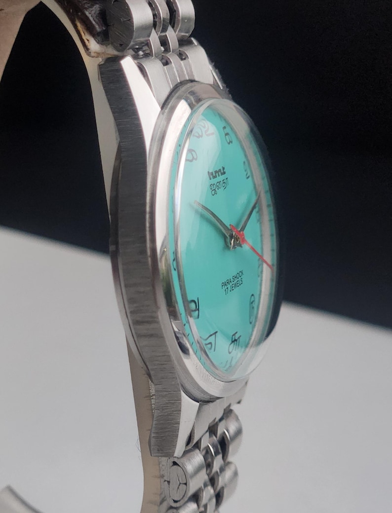 May include: A silver-toned wristwatch with a turquoise face and a matching band. The watch face features black numerals and hands, with a red second hand. The watch is labeled "PARA SHOCK 17 JEWELS".