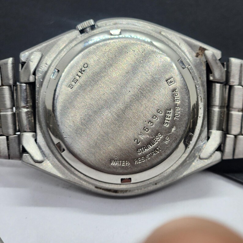 May include: Close-up of the back of a silver-toned Seiko watch. The watch has a stainless steel back with the brand name and model information. The watch band is also silver-toned and made of metal.