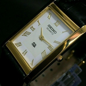 May include: A rectangular Seiko Quartz watch with a gold-tone case and a white face. The watch features gold Roman numeral hour markers and hands, and a black leather strap. The watch face displays the Seiko Quartz logo.