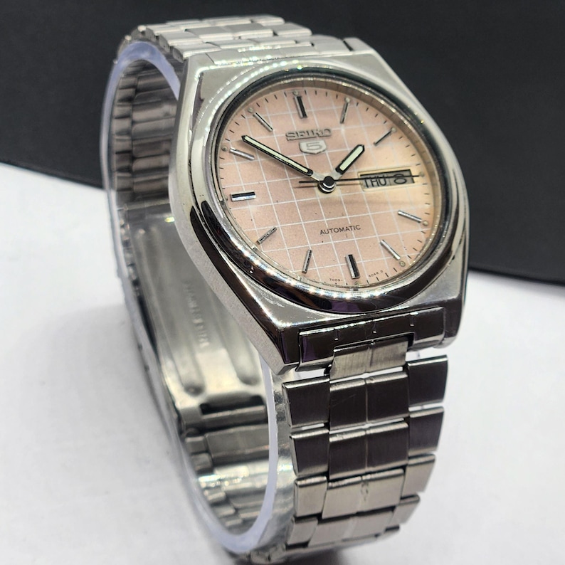 May include: Silver-toned Seiko 5 automatic watch with a light pink grid-patterned face. The watch features a day and date display and a stainless steel band. The watch is a classic timepiece.