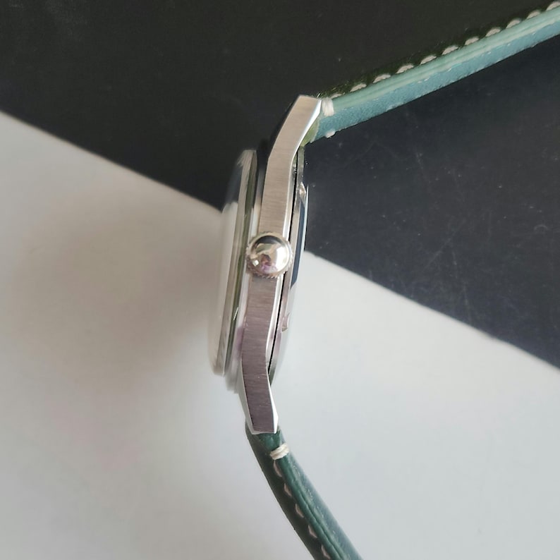 May include: A silver wristwatch with a round face and a light green leather band. The watch has a classic design with a small crown and a simple, elegant aesthetic. The band is stitched with white thread.