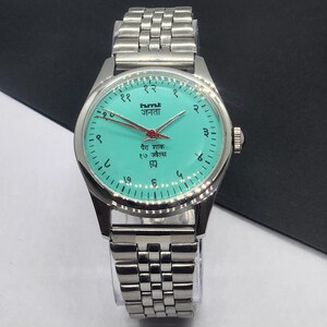May include: A silver-toned wristwatch with a turquoise face and a matching silver band. The watch face features black numerals, red hands, and text in a foreign language. The watch has a classic, vintage design.