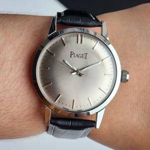 Puede incluir: Un reloj Piaget plateado con una correa de cuero negro. La esfera del reloj muestra el nombre de la marca y marcadores de hora minimalistas. El reloj est&aacute; en la mu&ntilde;eca de una persona.