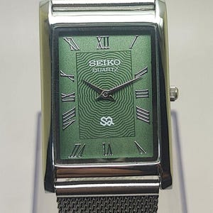 May include: A rectangular Seiko Quartz watch with a silver mesh band. The watch face is green with silver Roman numerals and hands. The watch face also has the Seiko Quartz logo and a unique design.