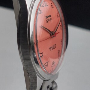 Puede incluir: Primer plano de un reloj de pulsera plateado con esfera color coral. El reloj presenta agujas negras y n&uacute;meros ar&aacute;bigos. La esfera del reloj incluye el texto "hmt" y "PARA SHOCK 17 JEWELS". La correa del reloj es de eslabones plateados.