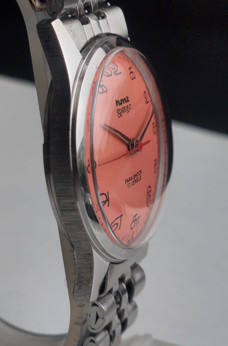 Puede incluir: Primer plano de un reloj de pulsera plateado con esfera color coral. El reloj presenta agujas negras y n&uacute;meros ar&aacute;bigos. La esfera del reloj incluye el texto "hmt" y "PARA SHOCK 17 JEWELS". La correa del reloj es de eslabones plateados.