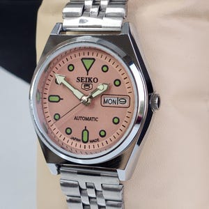 K&ouml;nnte beinhalten: Eine Seiko 5 Automatik-Uhr aus Edelstahl mit einem lachsfarbenen Zifferblatt. Die Uhr hat ein silbernes Armband, gr&uuml;ne Stundenmarkierungen und den Wochentag als "MON 9" angezeigt. Auf dem Zifferblatt steht au&szlig;erdem "SEIKO 5", "AUTOMATIC" und "JAPAN MADE".