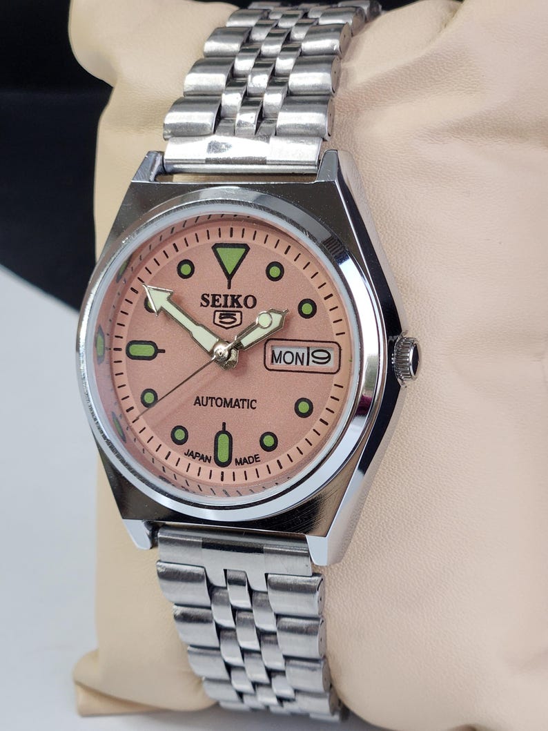 K&ouml;nnte beinhalten: Eine Seiko 5 Automatik-Uhr aus Edelstahl mit einem lachsfarbenen Zifferblatt. Die Uhr hat ein silbernes Armband, gr&uuml;ne Stundenmarkierungen und den Wochentag als "MON 9" angezeigt. Auf dem Zifferblatt steht au&szlig;erdem "SEIKO 5", "AUTOMATIC" und "JAPAN MADE".