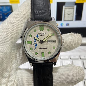 May include: A silver-toned Seiko automatic wristwatch with a black leather band. The watch face is off-white with green and blue accents, including the day and date display. The watch hands are luminous.