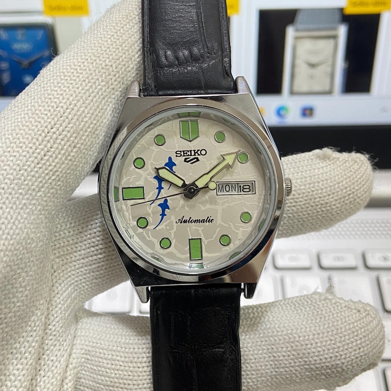 May include: A silver-toned Seiko automatic wristwatch with a black leather band. The watch face is off-white with green and blue accents, including the day and date display. The watch hands are luminous.
