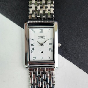 May include: A rectangular Seiko Quartz watch with a silver metal strap and case. The white watch face has black Roman numerals, black hands, and the Seiko logo. The watch strap is made of small, connected metal links.