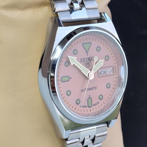 K&ouml;nnte beinhalten: Eine silberfarbene Seiko 5 Automatik-Armbanduhr mit lachsfarbenem Zifferblatt. Die Uhr verf&uuml;gt &uuml;ber ein Edelstahlarmband und eine Datumsanzeige. Das Zifferblatt hat Leuchtzeiger und -markierungen mit dem Text "SEIKO 5" und "AUTOMATIC".