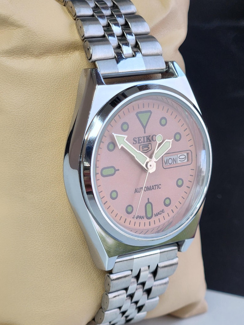 K&ouml;nnte beinhalten: Eine silberfarbene Seiko 5 Automatik-Armbanduhr mit lachsfarbenem Zifferblatt. Die Uhr verf&uuml;gt &uuml;ber ein Edelstahlarmband und eine Datumsanzeige. Das Zifferblatt hat Leuchtzeiger und -markierungen mit dem Text "SEIKO 5" und "AUTOMATIC".