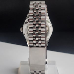 May include: A silver-toned wristwatch with a stainless steel band. The watch features a round face with visible internal mechanisms. The band is composed of small, rectangular links. The watch is displayed on a clear stand.