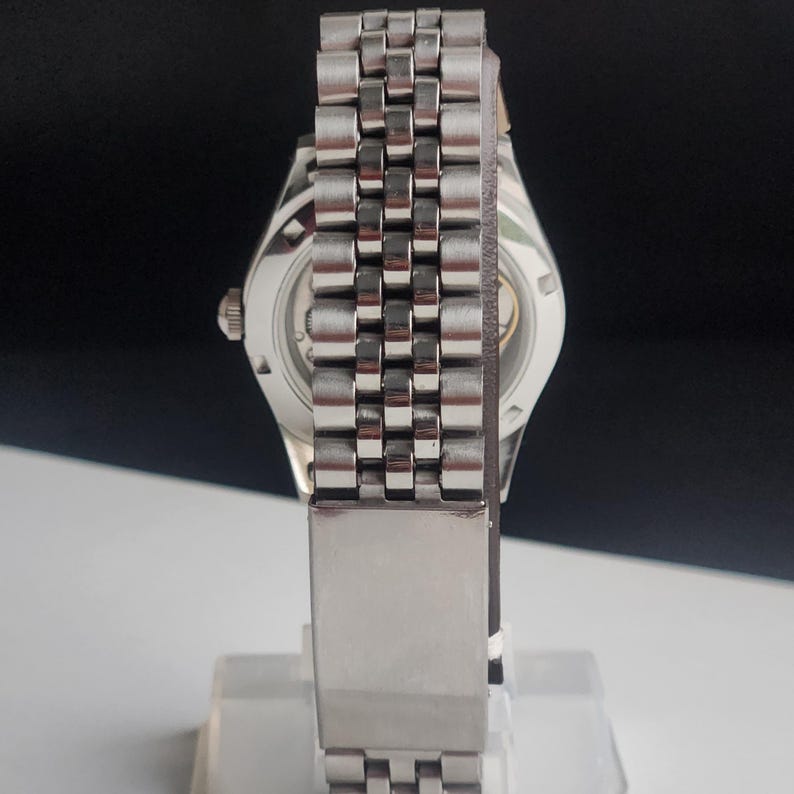 May include: A silver-toned wristwatch with a stainless steel band. The watch features a round face with visible internal mechanisms. The band is composed of small, rectangular links. The watch is displayed on a clear stand.
