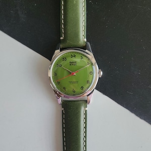 May include: A vintage wristwatch with a green face and a matching green leather band. The watch has silver-colored casing and hands, with Arabic numerals and text in a foreign language. The band is stitched with white thread.