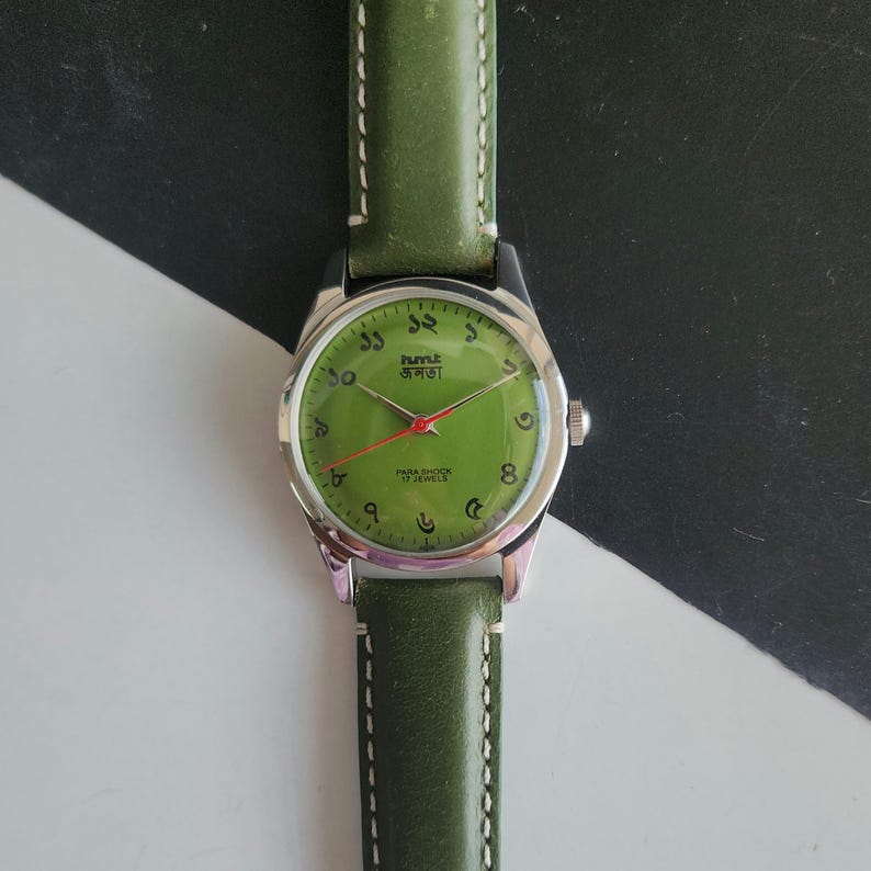 May include: A vintage wristwatch with a green face and a matching green leather band. The watch has silver-colored casing and hands, with Arabic numerals and text in a foreign language. The band is stitched with white thread.